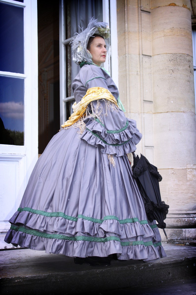 May include: A woman in a gray dress with green trim and a white bonnet stands in front of a white door. She is holding a black umbrella.