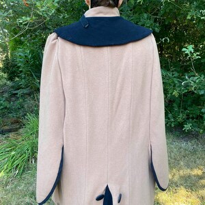 May include: A tan wool coat with a black collar and trim. The coat has a long, flowing silhouette and a single button closure at the back.