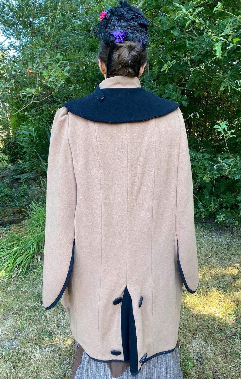 May include: A tan wool coat with a black collar and trim. The coat has a long, flowing silhouette and a single button closure at the back.