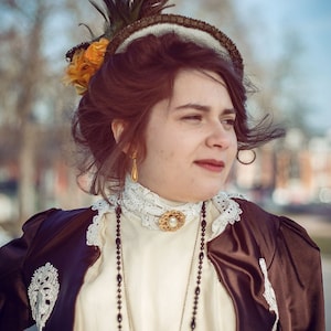 Puede incluir: Un atuendo de estilo vintage. Chaqueta de satén marrón sobre un vestido crema con ribete de encaje. Collar de cuentas, broche dorado y sombrero adornado con plumas y flores. La estética general recuerda a la época victoriana. Moda histórica.