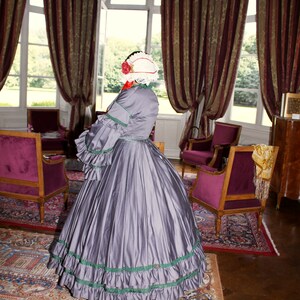 May include: A woman in a gray dress with a white and red hat stands in a room with a patterned rug and antique furniture. The dress has green trim and a ruffled skirt.