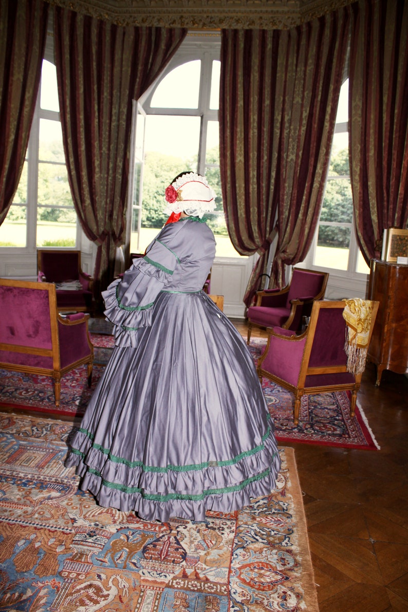 May include: A woman in a gray dress with a white and red hat stands in a room with a patterned rug and antique furniture. The dress has green trim and a ruffled skirt.