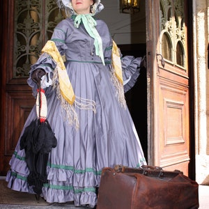 May include: A woman in a gray dress with a yellow and white scarf and a hat is standing in a doorway. She is holding a black umbrella and a brown leather suitcase.