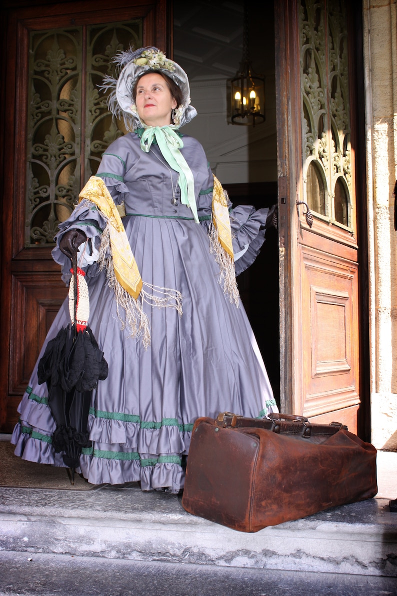 May include: A woman in a gray dress with a yellow and white scarf and a hat is standing in a doorway. She is holding a black umbrella and a brown leather suitcase.