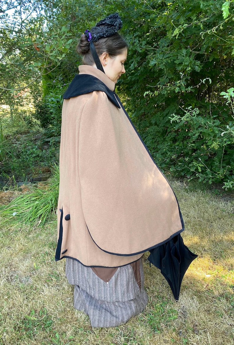 May include: A brown cape with a black velvet collar and black trim. The cape is worn over a striped skirt and a black umbrella is held in the right hand.