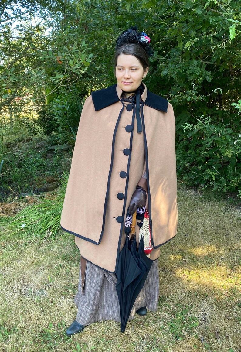 May include: A woman wearing a brown cape with black trim and a black ribbon around her neck. She is holding a black umbrella and a pair of gloves.