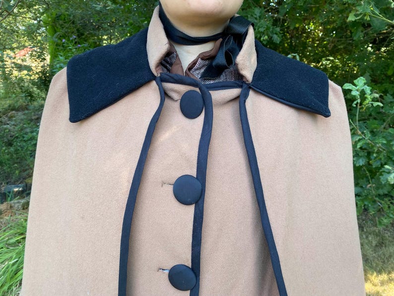 May include: A tan wool cape with a black velvet collar and black trim. The cape has four black buttons down the front.