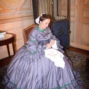 May include: A woman in a gray dress with green trim sits in a chair and works on an embroidery project. She is sitting in a room with a patterned rug and a wooden chair.