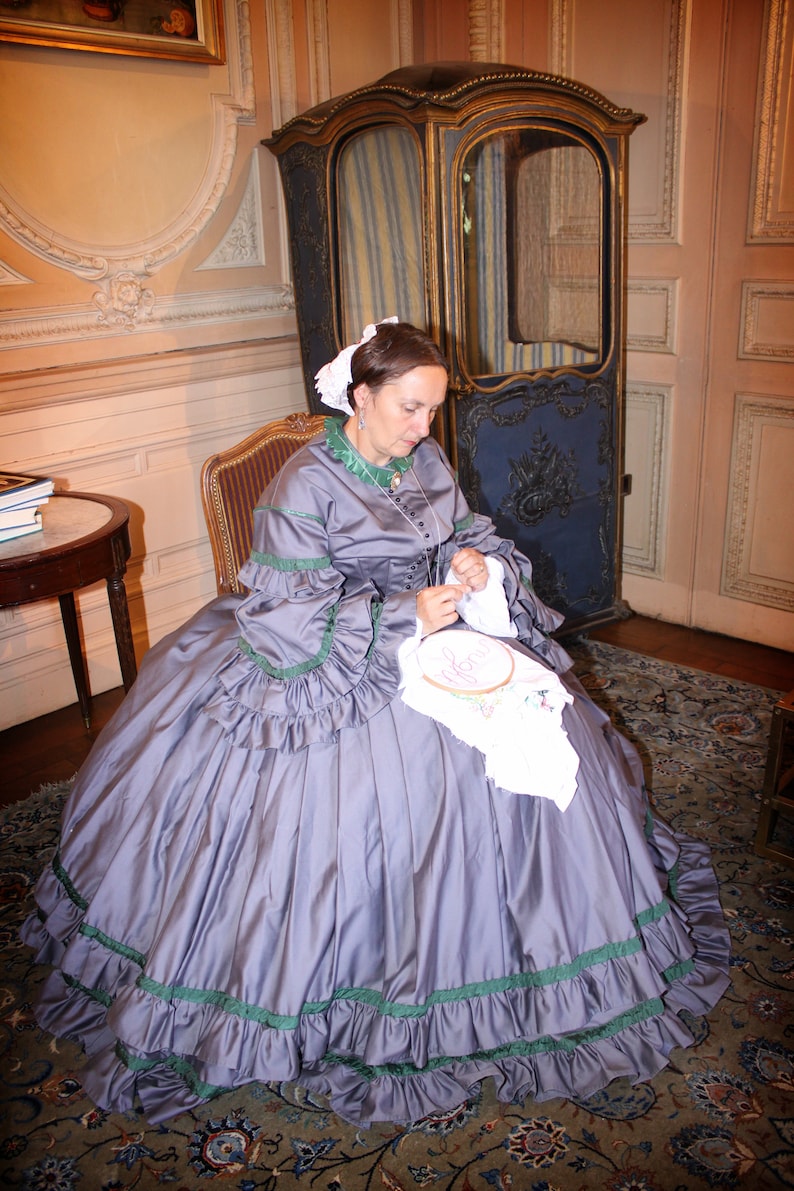 May include: A woman in a gray dress with green trim sits in a chair and works on an embroidery project. She is sitting in a room with a patterned rug and a wooden chair.