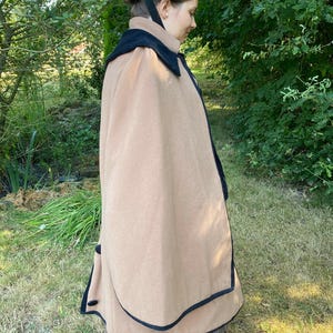May include: A tan cape with a black trim and a black velvet collar. The cape is worn over a brown and white striped skirt.