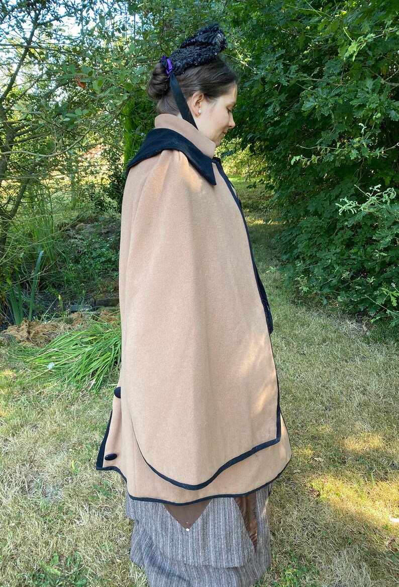 May include: A tan cape with a black trim and a black velvet collar. The cape is worn over a brown and white striped skirt.