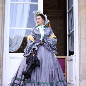 May include: A woman in a gray dress with green and yellow trim stands in front of a white door. She is wearing a hat with a green ribbon and holding a black umbrella.