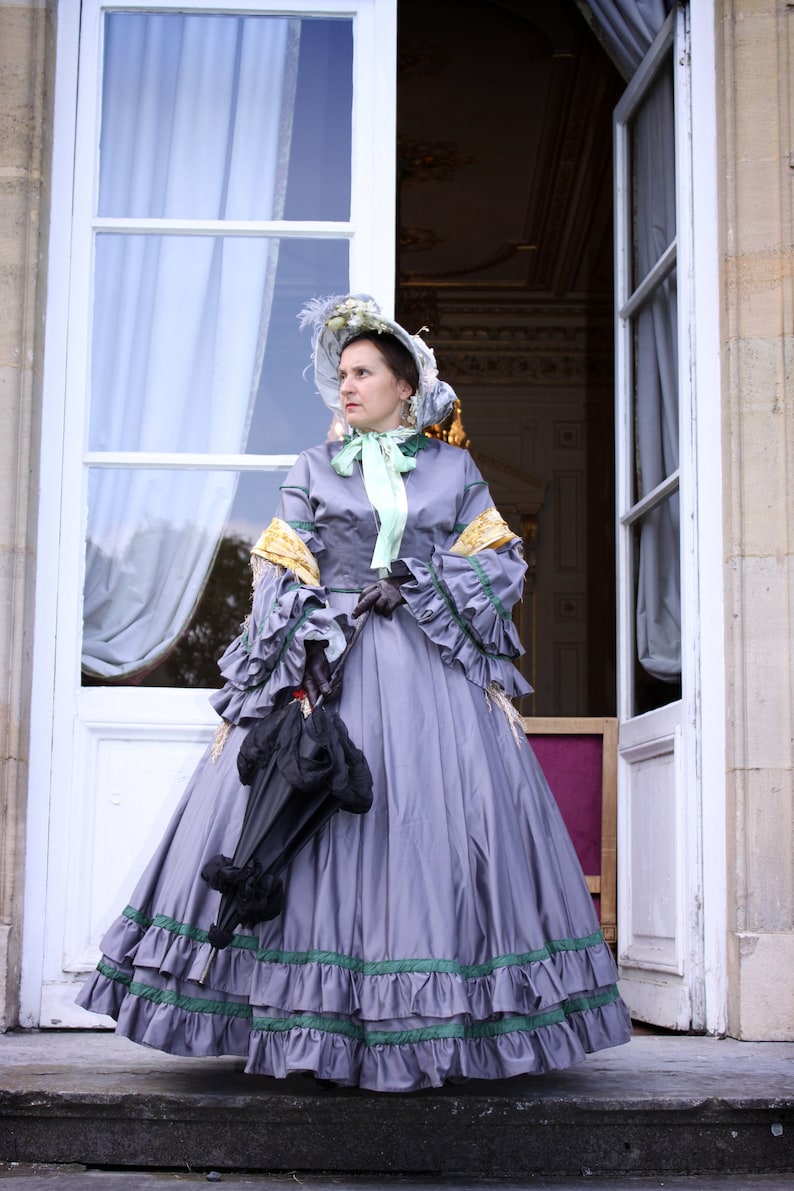 May include: A woman in a gray dress with green and yellow trim stands in front of a white door. She is wearing a hat with a green ribbon and holding a black umbrella.