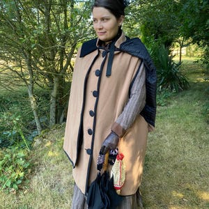 May include: A woman wearing a brown cape with black trim and a black umbrella. She is wearing a brown and white striped dress and black gloves.