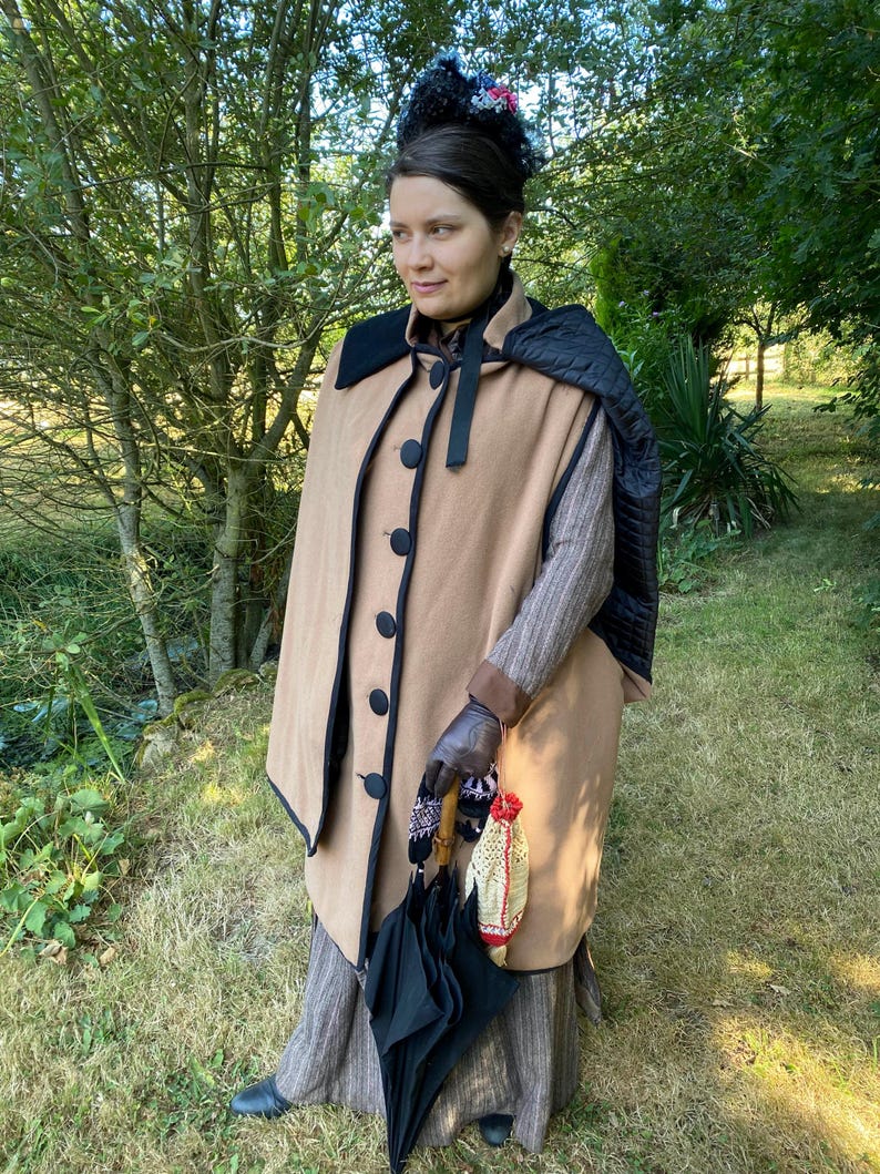 May include: A woman wearing a brown cape with black trim and a black umbrella. She is wearing a brown and white striped dress and black gloves.
