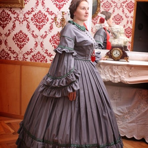 May include: A woman in a gray dress with green trim and ruffles stands in front of a mirror. The dress has a fitted bodice and a full skirt. The woman is wearing her hair up and has a necklace on.