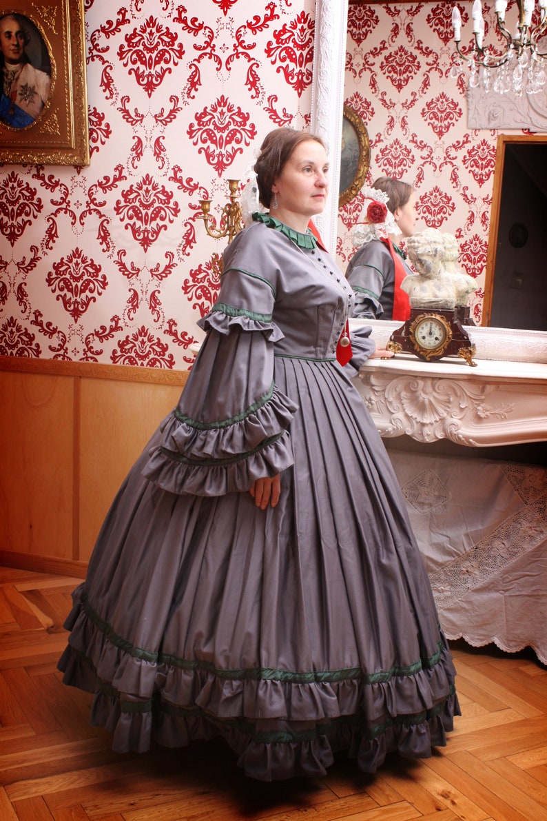 May include: A woman in a gray dress with green trim and ruffles stands in front of a mirror. The dress has a fitted bodice and a full skirt. The woman is wearing her hair up and has a necklace on.
