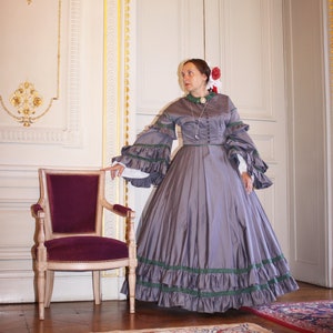 May include: A woman in a gray, floor-length dress with green trim and a white lace collar stands in a room with a patterned rug and a wooden chair. She is wearing a red flower in her hair.