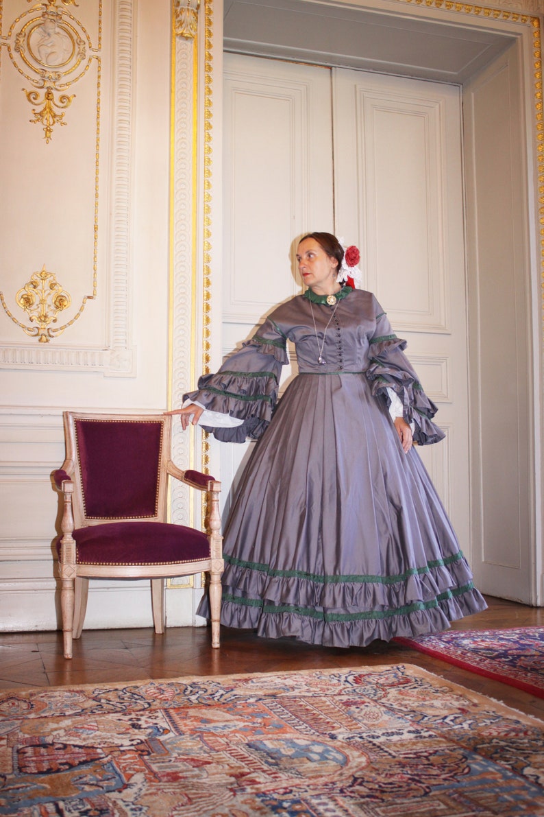 May include: A woman in a gray, floor-length dress with green trim and a white lace collar stands in a room with a patterned rug and a wooden chair. She is wearing a red flower in her hair.