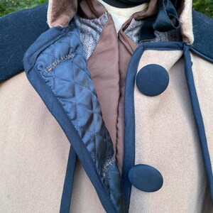 May include: A beige wool cape with a black velvet collar and trim. The cape has two black buttons on the front.