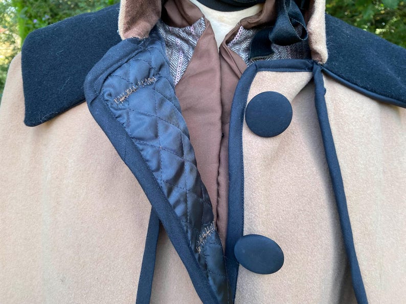 May include: A beige wool cape with a black velvet collar and trim. The cape has two black buttons on the front.