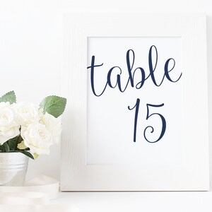 Può includere: Cornice bianca con sfondo bianco e testo blu scuro che recita "tavolo 15". La cornice è appoggiata su una superficie bianca con un secchiello bianco di rose bianche di fronte.