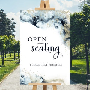 May include: A white sign on a wooden easel with the words "OPEN seating PLEASE SEAT YOURSELF" in black script. The sign has a watercolor design with blue, black, and gold accents. The easel is outdoors in a park setting.