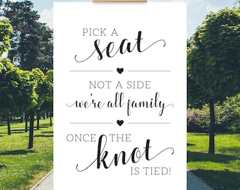 May include: A white sign on a wooden easel with the text "PICK A seat, NOT A SIDE, we're all family, ONCE THE knot IS TIED!" in black script. The sign is displayed outdoors with trees and a pathway in the background.