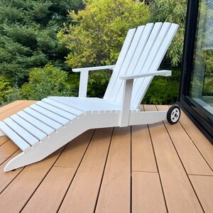 K&ouml;nnte beinhalten: Wei&szlig;er Adirondack-Stuhl aus Holz mit Fu&szlig;st&uuml;tze und R&auml;dern. Der Stuhl steht auf einem Holzdeck.