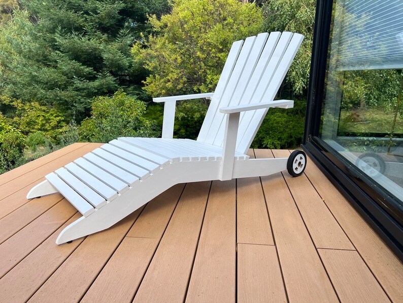 K&ouml;nnte beinhalten: Wei&szlig;er Adirondack-Stuhl aus Holz mit Fu&szlig;st&uuml;tze und R&auml;dern. Der Stuhl steht auf einem Holzdeck.