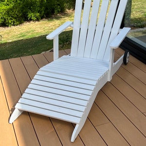 K&ouml;nnte beinhalten: Wei&szlig;er Holz-Adirondack-Stuhl mit Fu&szlig;st&uuml;tze. Der Stuhl steht auf einem Holzdeck.