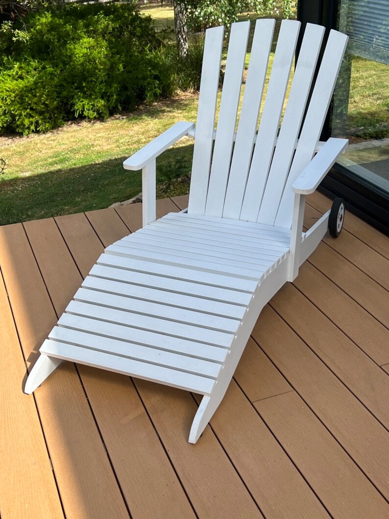 K&ouml;nnte beinhalten: Wei&szlig;er Holz-Adirondack-Stuhl mit Fu&szlig;st&uuml;tze. Der Stuhl steht auf einem Holzdeck.