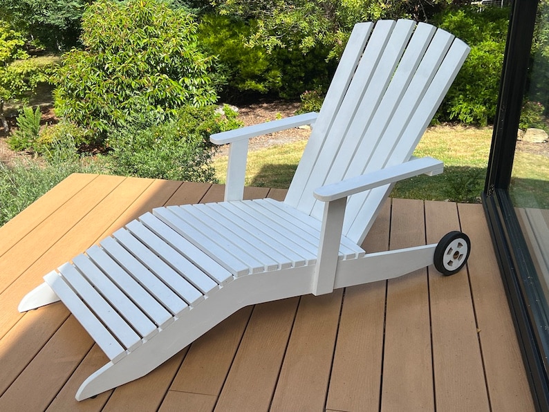 K&ouml;nnte beinhalten: Wei&szlig;er Adirondack-Stuhl aus Holz mit Fu&szlig;st&uuml;tze und einem Rad auf einer Seite. Der Stuhl steht auf einem Holzdeck.