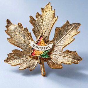 May include: A gold-toned metal brooch shaped like a large maple leaf with a textured surface. Centered on the leaf is a smaller, colorful enamel maple leaf in red, yellow, and green, featuring a white banner with the text MONTREAL.