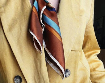 Può includere: Blazer giallo senape con design doppiopetto. Una sciarpa di seta a righe marroni, blu e arancioni è annodata al collo. È visibile una collana di perle. Il blazer ha due bottoni visibili.