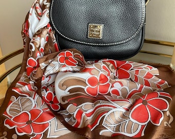 Könnte beinhalten: Eine schwarze Leder-Umhängetasche mit goldenem Dooney & Bourke-Logo auf einem braunen und weißen floralen Schal.