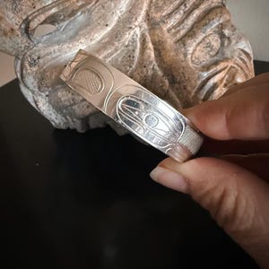 Op de afbeelding: Een zilveren armband met ingewikkelde gegraveerde ontwerpen. De armband heeft een reeks gestileerde vormen en patronen, mogelijk een cultureel motief. Het metaal heeft een gepolijste afwerking die licht reflecteert. De armband wordt tegen een neutrale achtergrond gehouden.