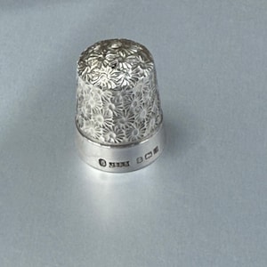 May include: A silver thimble featuring a detailed embossed floral pattern covering the dome. The smooth base band displays a series of small stamped hallmarks, including the number 6, indicating its specific production and metal quality.