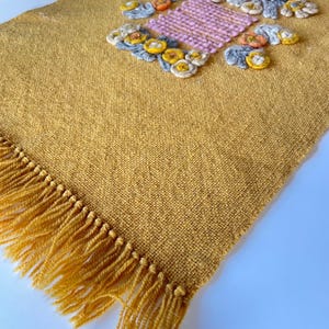 Puede incluir: Camino de mesa tejido amarillo mostaza con borde de flecos. El camino de mesa presenta diseños florales bordados en rosa, amarillo, gris y naranja. La sección bordada mide aproximadamente 30 cm de ancho.
