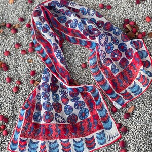 May include: A white silk scarf with a red, blue, and white geometric pattern. The scarf is folded and laid out on a bed of small gray stones. The scarf has the word "Usnao" printed on the bottom right corner.
