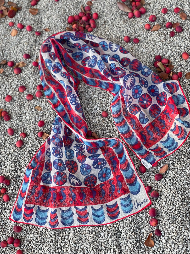 May include: A white silk scarf with a red, blue, and white geometric pattern. The scarf is folded and laid out on a bed of small gray stones. The scarf has the word "Usnao" printed on the bottom right corner.