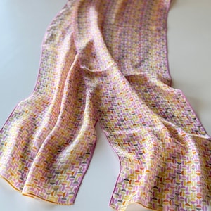 Peut inclure: Une longue écharpe rectangulaire avec un motif répétitif de demi-cercles colorés dans les tons de rose, jaune, orange et violet. L'écharpe a une texture soyeuse et un drapé délicat et fluide. Les bords sont finis avec une fine bordure orange.