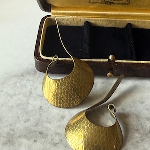 May include: Gold-coloured earrings with a textured, honeycomb pattern. The earrings have a distinctive, curved design and are shown on a marble surface near a vintage jewellery box.