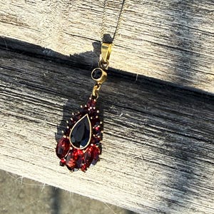 May include: A gold-toned necklace with a teardrop pendant. The pendant features a large, black, teardrop-shaped stone surrounded by smaller, red stones. The necklace is displayed on a weathered wooden surface.