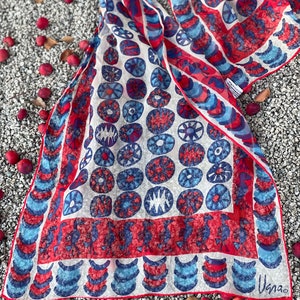 May include: A white silk scarf with a red and blue geometric pattern. The pattern features circles, crescents, and stylized birds. The scarf is signed "Usna" in the bottom right corner.