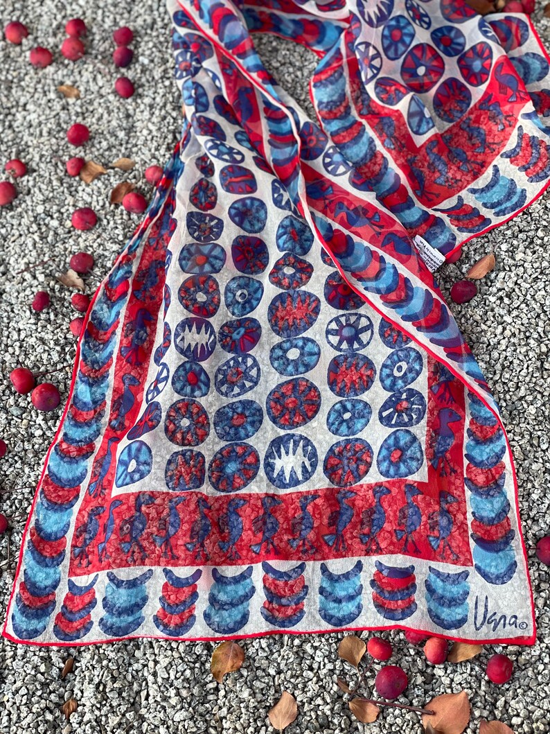 May include: A white silk scarf with a red and blue geometric pattern. The pattern features circles, crescents, and stylized birds. The scarf is signed "Usna" in the bottom right corner.