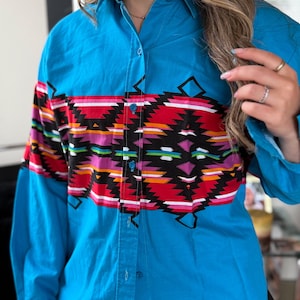 Pode incluir: Uma camisa turquesa com botões e um padrão geométrico colorido no peito. O padrão inclui formas vermelhas, pretas, brancas, rosa e laranja. A camisa tem mangas compridas e uma gola clássica.