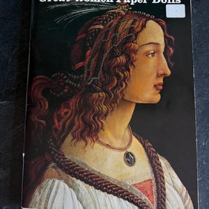 May include: A vintage book titled "Great Women Paper Dolls" featuring a Renaissance-style portrait of a woman with auburn hair and a decorative necklace. The cover has a dark background and the title is in white text.