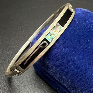Peut inclure: Bracelet jonc articulé argenté avec incrustations noires et accents de coquillage d'ormeau irisé. Le bracelet présente un panneau noir rectangulaire et un élément de design incurvé. Le bracelet repose sur une surface en velours bleu.