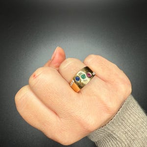 Op de afbeelding: Een gouden ring met drie ingezette edelstenen: blauw, groen en rood. De ring heeft een brede band en kleine diamanten aan weerszijden van de edelstenen. De ring wordt op een hand getoond.